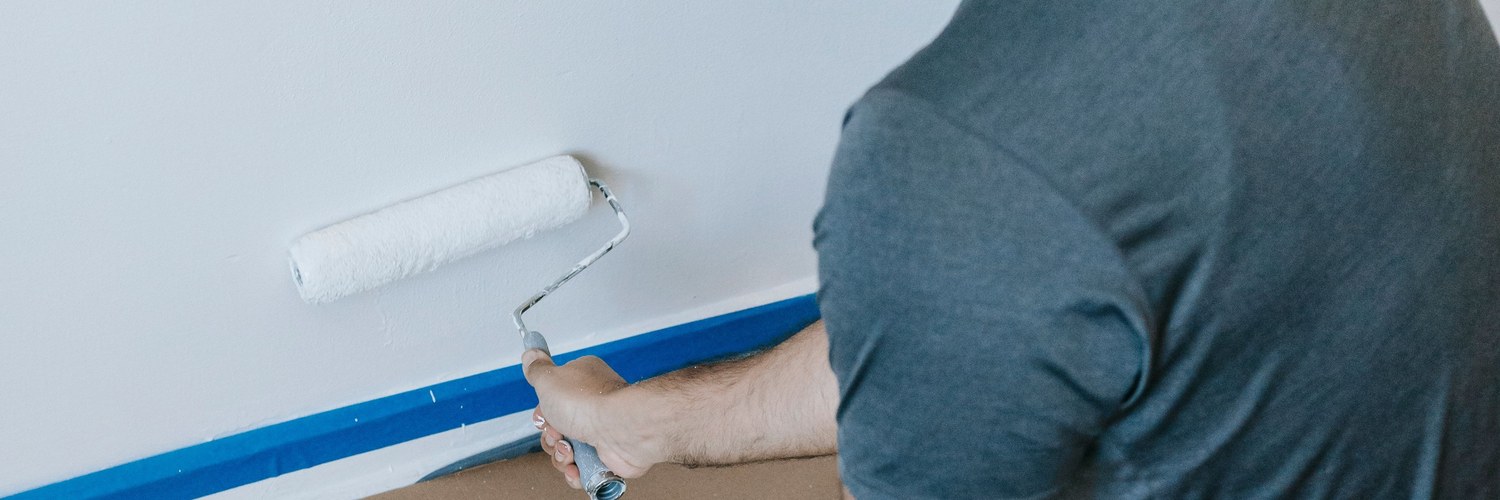 Painter rolling white paint onto a wall, blue tape along the baseboard
